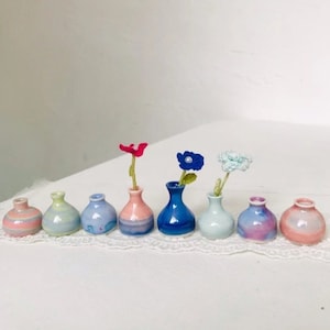May include: A collection of eight small, ceramic vases in various pastel colors, each with a unique shape and design. Some vases hold small, handmade flowers. The vases are arranged in a row on a white lace surface.
