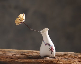 May include: A small white ceramic vase with a floral design, featuring a single dried flower with a yellow center.
