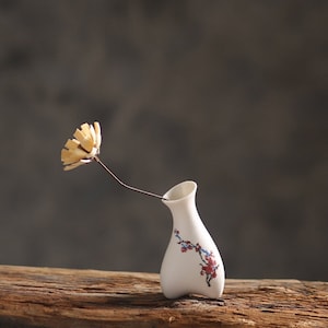 Pode incluir: Um pequeno vaso de cerâmica branco com um design floral, apresentando uma única flor seca com um centro amarelo.