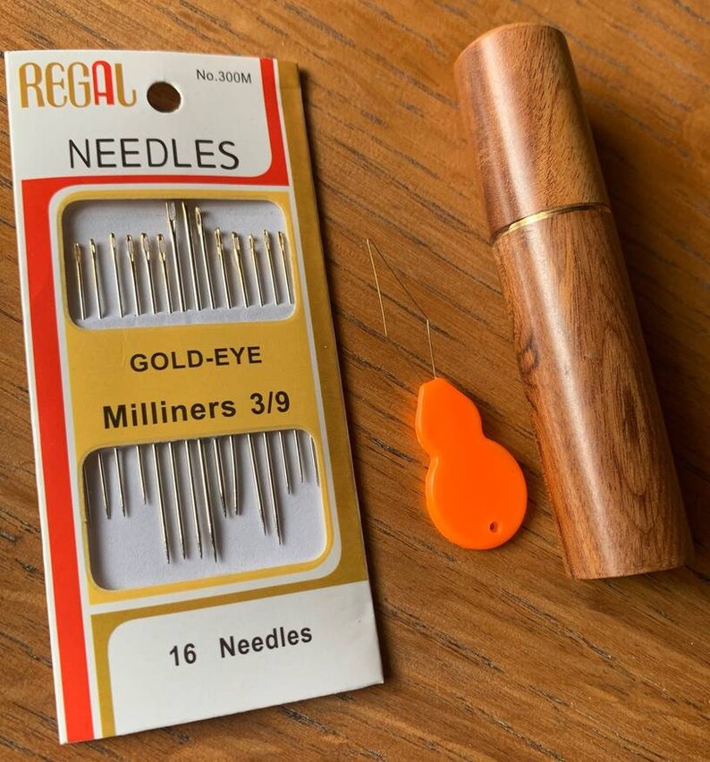 May include: A package of 16 gold-eye milliners needles, size 3/9, in a white and yellow box with the brand name "REGAL" and the number "300M". The needles are packaged with a wooden case and a small orange needle threader.