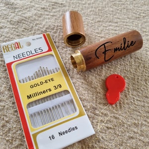 May include: A wooden needle case with a brass top and a gold-eye needle threader. The case is engraved with the name "Emilie". The case is open and a package of 16 gold-eye milliners needles is visible. The package is red and white with the text "REGAL NEEDLES No. 300M GOLD-EYE Milliners 3/9 16 Needles".