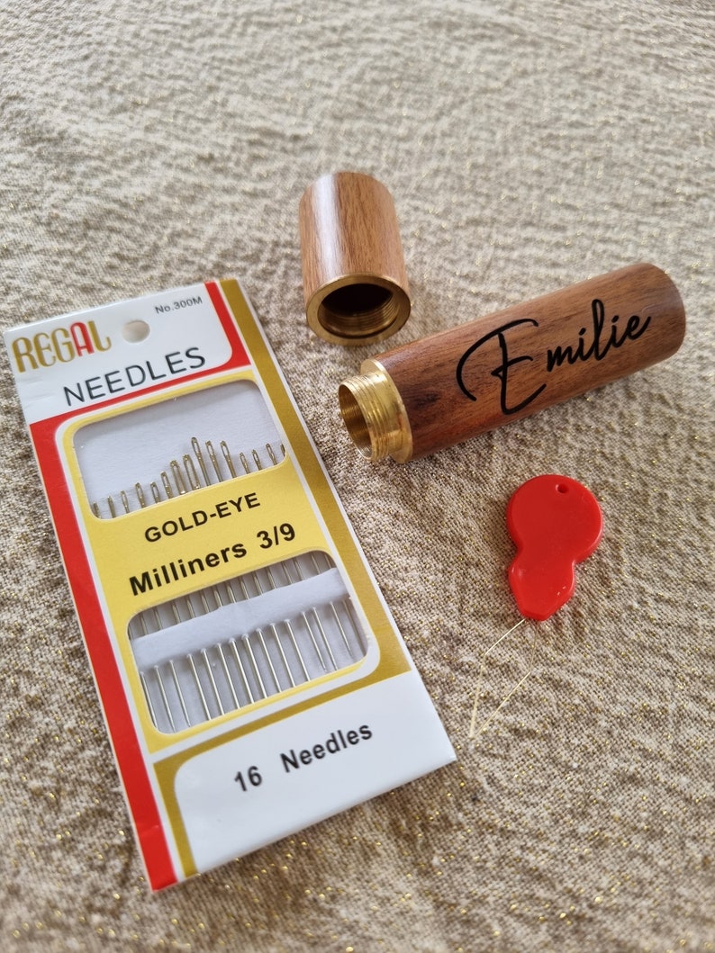 May include: A wooden needle case with a brass top and a gold-eye needle threader. The case is engraved with the name "Emilie". The case is open and a package of 16 gold-eye milliners needles is visible. The package is red and white with the text "REGAL NEEDLES No. 300M GOLD-EYE Milliners 3/9 16 Needles".