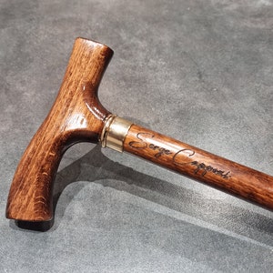 Può includere: Un bastone da passeggio in legno marrone con un manico curvo e una fascia in ottone. Il bastone è inciso con il nome "Serge Capponi".