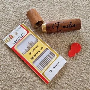 May include: A wooden needle case with the name "Emilie" engraved on it. The case is open and a package of 16 gold-eye milliners needles is visible. The package is red and white with the brand name "Regal" printed on it.