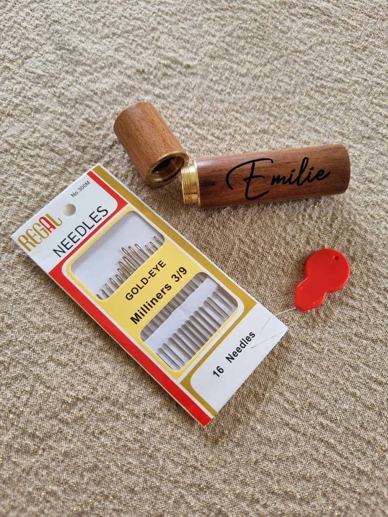 May include: A wooden needle case with the name "Emilie" engraved on it. The case is open and a package of 16 gold-eye milliners needles is visible. The package is red and white with the brand name "Regal" printed on it.