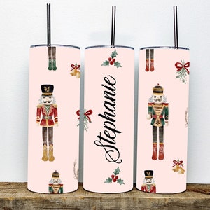 May include: Three pink stainless steel tumblers with a Christmas nutcracker design. The tumblers feature a watercolor illustration of a nutcracker, holly, bows, and bells. The middle tumbler has the name "Stephanie" written on it.