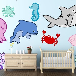 May include: A nursery scene featuring cartoon sea creatures: an octopus, seahorse, dolphin, shark, crab, and seashell. The room includes a light-colored wooden dresser, a crib, and decorative coral. The walls are painted a light blue.