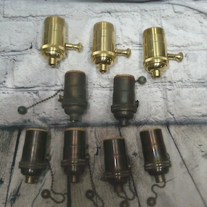 May include: A collection of nine light bulb sockets arranged in three rows. The top row features three polished brass sockets with side turn knobs. The middle and bottom rows contain six dark, aged bronze-toned sockets with pull chain switches.
