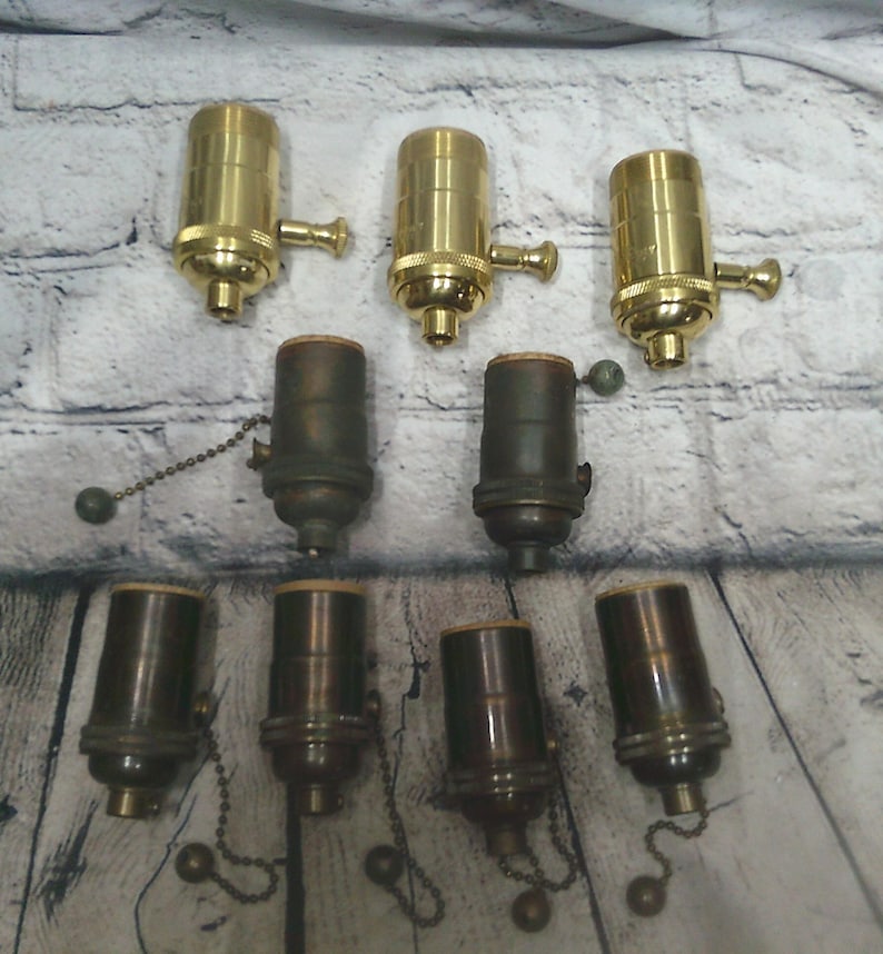 May include: A collection of nine light bulb sockets arranged in three rows. The top row features three polished brass sockets with side turn knobs. The middle and bottom rows contain six dark, aged bronze-toned sockets with pull chain switches.