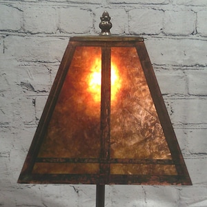 May include: A trapezoidal lamp shade with a warm, amber-toned mottled surface and dark brown structural framing. The shade features a decorative metal finial at the top center and emits a soft, glowing light from within the translucent panels.