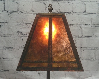 Può includere: Un paralume trapezoidale con una superficie screziata dai caldi toni ambrati e una cornice strutturale marrone scuro. Il paralume presenta un terminale decorativo in metallo al centro della parte superiore ed emette una luce soffusa.