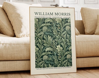 May include: A framed art print featuring a dark green and cream floral pattern. The print is titled "William Morris" and includes text about an exhibition. The artwork is displayed on a beige sofa in a well-lit room with wooden flooring.