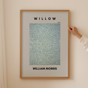 Könnte beinhalten: Ein gerahmter Kunstdruck mit einem William Morris-Design, betitelt "Willow" von 1874. Das Kunstwerk zeigt ein sich wiederholendes Muster aus stilisierten grünen Blättern auf hellblauem Hintergrund. Der Rahmen ist aus hellbraunem Holz.