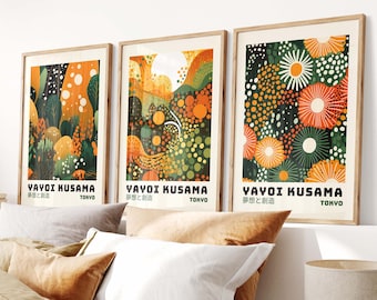 May include: Three framed art prints with a green and orange abstract floral design. The prints feature the text "YAYOI KUSAMA" and "夢と創造" in Japanese. The prints are hanging on a white wall above a bed with brown bedding.