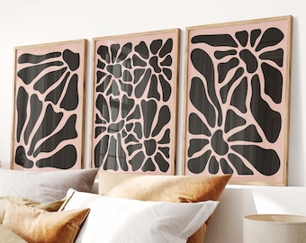 May include: Three abstract art prints with black and white floral designs on a light pink background. The prints are framed in wood and are hanging on a white wall above a bed with brown linen bedding.
