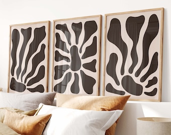 May include: Three framed abstract art prints with black and white organic shapes on a light beige background. The prints are hanging on a white wall above a bed with brown and white bedding.