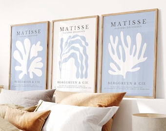 May include: Three framed prints of Matisse's "Papiers Decoupes" artwork. The prints are on a light blue background with white cut-out designs of stylized plants. The prints are hung on a white wall above a bed with brown and white bedding.