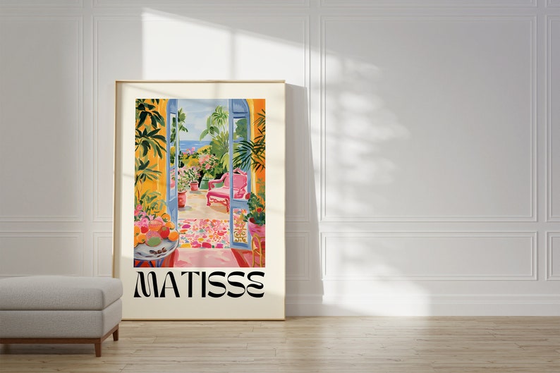 Framed poster featuring a colorful Matisse-inspired painting of an open balcony with lush plants, pink chair, and a view of the sea. Bright colors and floral patterns stand out against a white wall in a sunlit modern interior.
