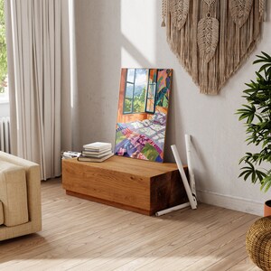 May include: A wooden bench with a stack of books on top. The bench is in a room with a window and a painting of a bedroom with a colorful quilt on the bed.