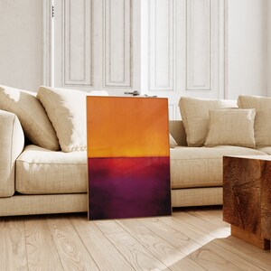 May include: An abstract painting with a gold frame, featuring a gradient of orange and purple hues. The artwork is displayed on a wooden floor, leaning against a beige sofa in a well-lit room. The painting's colors evoke a sunset.