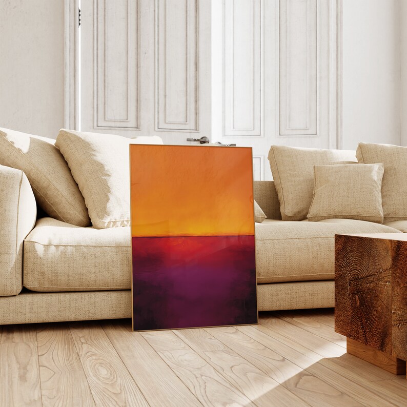 May include: An abstract painting with a gold frame, featuring a gradient of orange and purple hues. The artwork is displayed on a wooden floor, leaning against a beige sofa in a well-lit room. The painting's colors evoke a sunset.