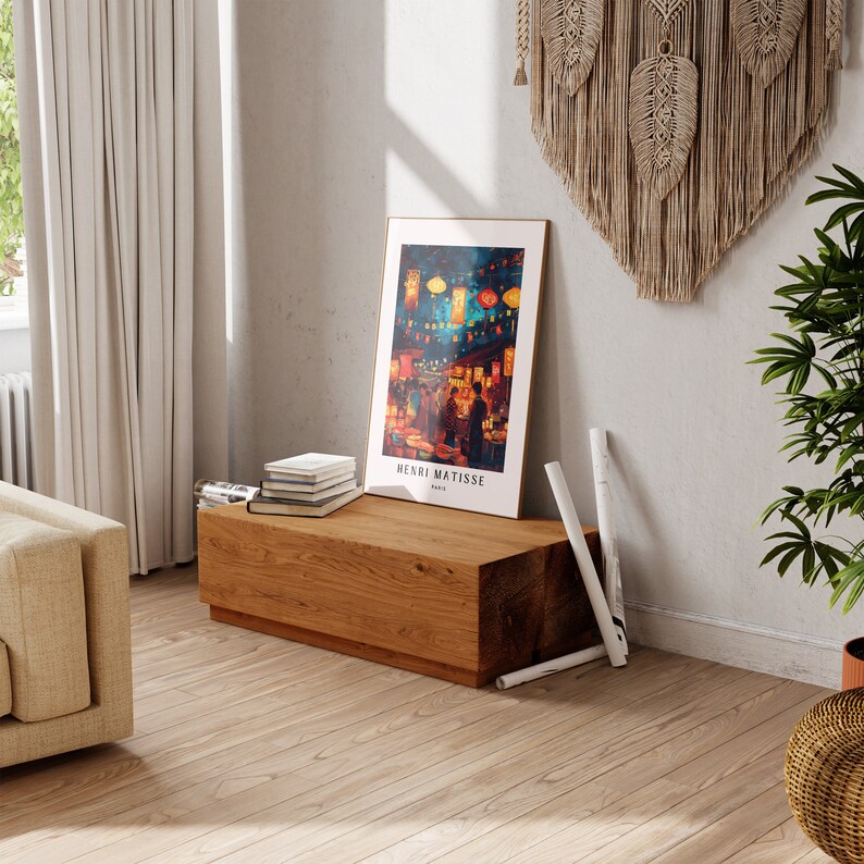 May include: A wooden bench with a framed print of a painting by Henri Matisse. The print depicts a night scene with colourful lanterns and people walking in a street. The bench is decorated with a stack of books and rolled up newspapers.