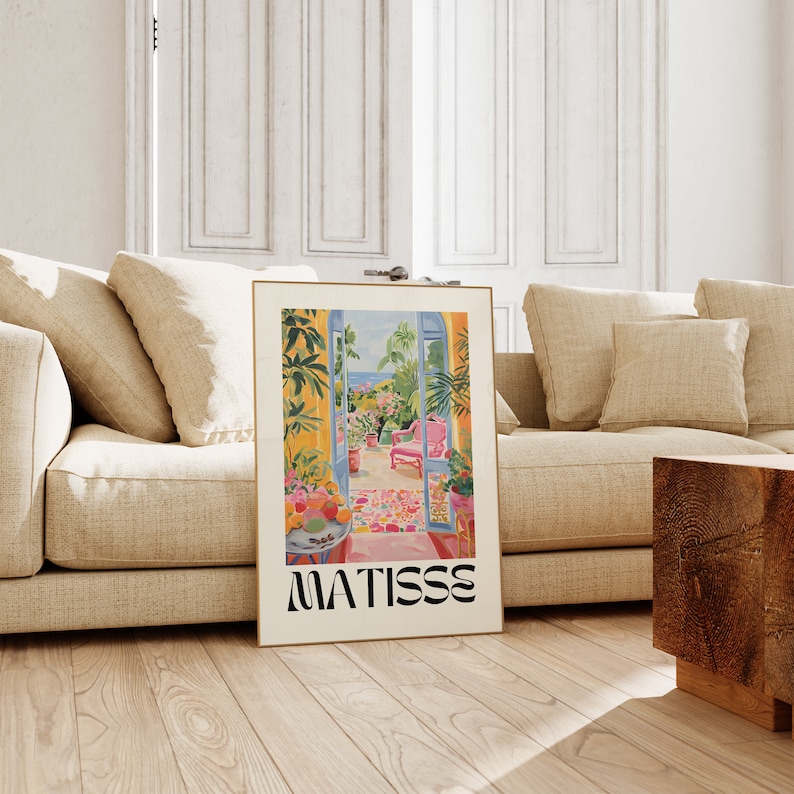 Framed Matisse-inspired poster leaning against a beige sofa in a modern living room. The vibrant artwork depicts a balcony garden scene with lush plants and a pink chair, set in a minimalist space with neutral tones and wooden flooring.