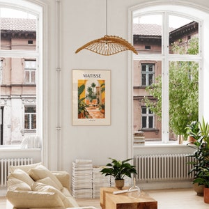 May include: A living room interior with a white wall, a large window, a beige couch, a stack of books, a potted plant, a wooden coffee table, and a poster with the text "MATISSE" and a colorful illustration of a garden.