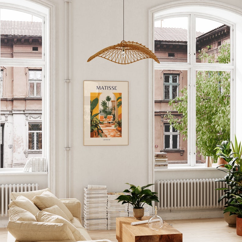 May include: A living room interior with a white wall, a large window, a beige couch, a stack of books, a potted plant, a wooden coffee table, and a poster with the text "MATISSE" and a colorful illustration of a garden.
