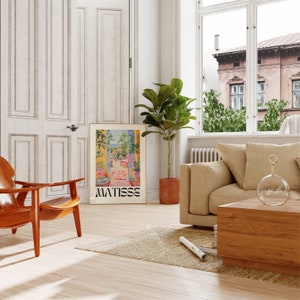 Framed Matisse-inspired poster leaning against a wall in a bright living room. The scene includes a beige sofa, wooden chair, potted plant, and large windows with views of an exterior building. The room has a cozy, natural aesthetic with warm tones.