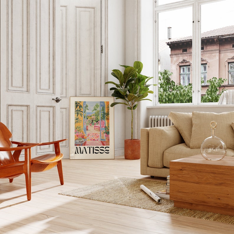 Framed Matisse-inspired poster leaning against a wall in a bright living room. The scene includes a beige sofa, wooden chair, potted plant, and large windows with views of an exterior building. The room has a cozy, natural aesthetic with warm tones.