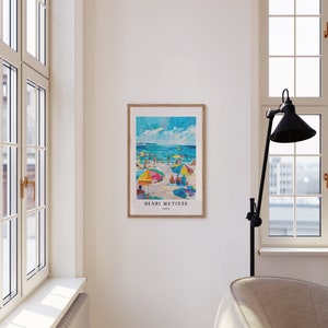 May include: A framed print of a painting by Henri Matisse depicting a beach scene with colorful umbrellas and people enjoying the sun and water. The print is in a wooden frame and has the artist's name and "Paris" printed below the image.