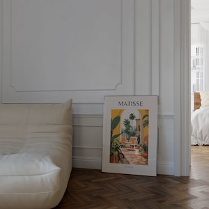 May include: A white couch with a white throw pillow sits in front of a white wall with a framed poster that says "MATISSE" and features a colorful illustration of an archway with palm trees and a patio scene.