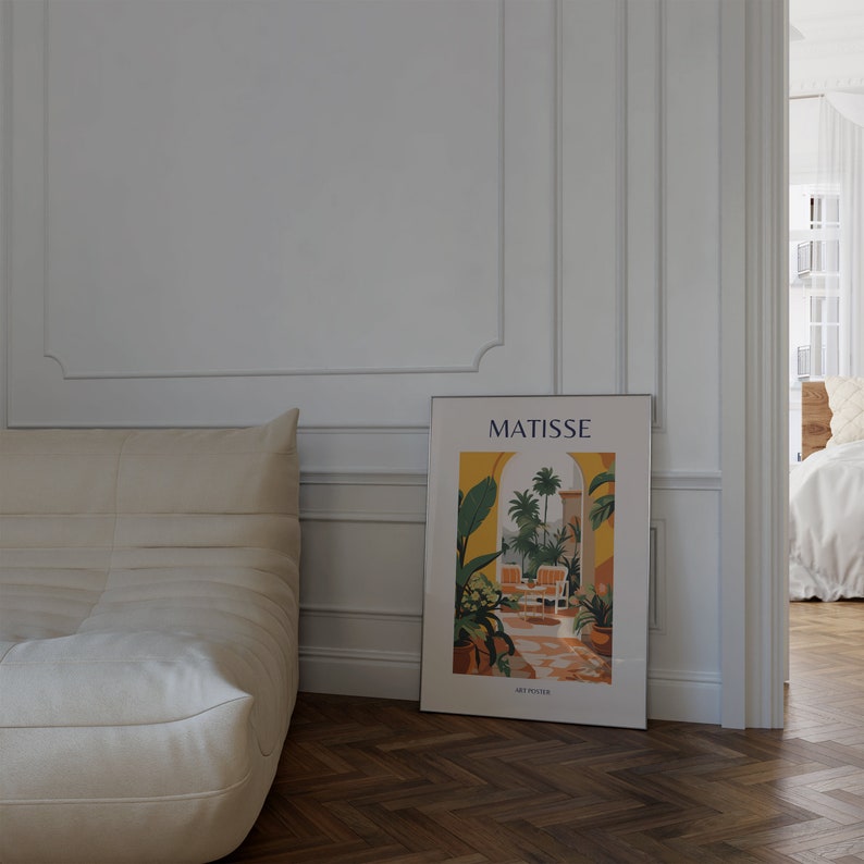 May include: A white couch with a white throw pillow sits in front of a white wall with a framed poster that says "MATISSE" and features a colorful illustration of an archway with palm trees and a patio scene.