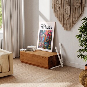 May include: A wooden bench with a framed print of a colorful painting featuring birds and foliage. The print has the text "Henri Matisse" at the top. The bench is in a room with a white wall and a macrame wall hanging.