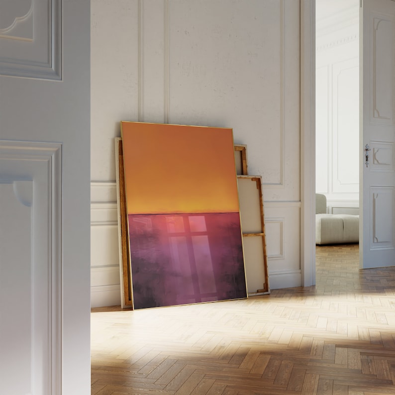 May include: An abstract art print featuring a gradient of orange and purple hues, leaning against a white wall. The artwork has a gold frame and is displayed in a room with a wooden floor.