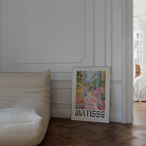 Framed Matisse-inspired poster leaning against a white paneled wall beside a beige sofa. The vibrant artwork shows a colorful garden balcony scene. The dark herringbone floor and soft lighting create a cozy, sophisticated atmosphere.