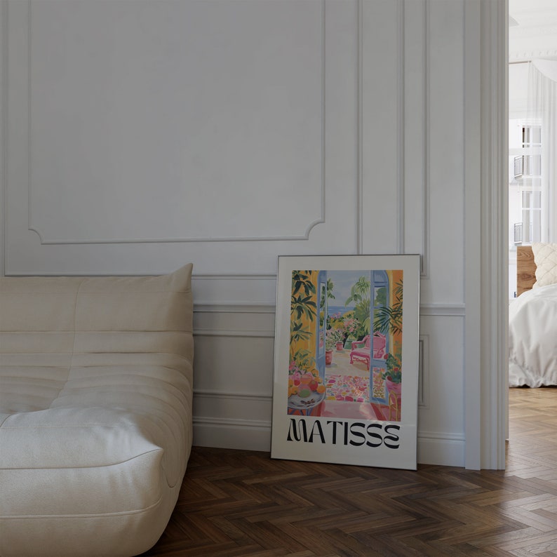 Framed Matisse-inspired poster leaning against a white paneled wall beside a beige sofa. The vibrant artwork shows a colorful garden balcony scene. The dark herringbone floor and soft lighting create a cozy, sophisticated atmosphere.