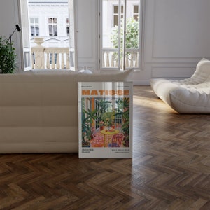 May include: A poster featuring a painting by Henri Matisse titled "The Garden." The poster is framed and has a white background with the artist's name and the title of the painting in bold, red letters. The painting depicts a colorful garden scene with a table and chairs.