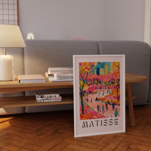 May include: A framed art print depicting a vibrant park scene with figures and trees in pinks, oranges, and greens. The print is labelled "MATISSE" in black at the bottom. The artwork is displayed on a wooden floor.