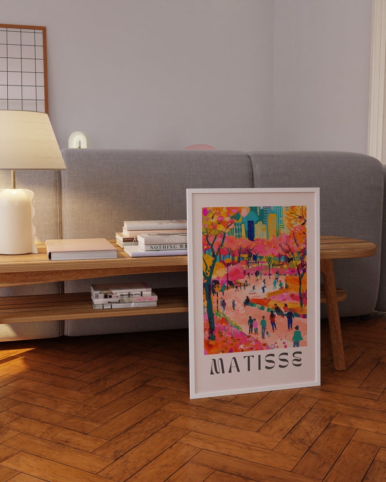 May include: A framed art print depicting a vibrant park scene with figures and trees in pinks, oranges, and greens. The print is labelled "MATISSE" in black at the bottom. The artwork is displayed on a wooden floor.
