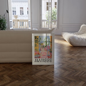 Framed Matisse-inspired poster propped against a beige sofa in a bright room with large open windows and a balcony view. The modern interior features herringbone wood flooring and soft, curved furniture, creating an elegant, airy feel.