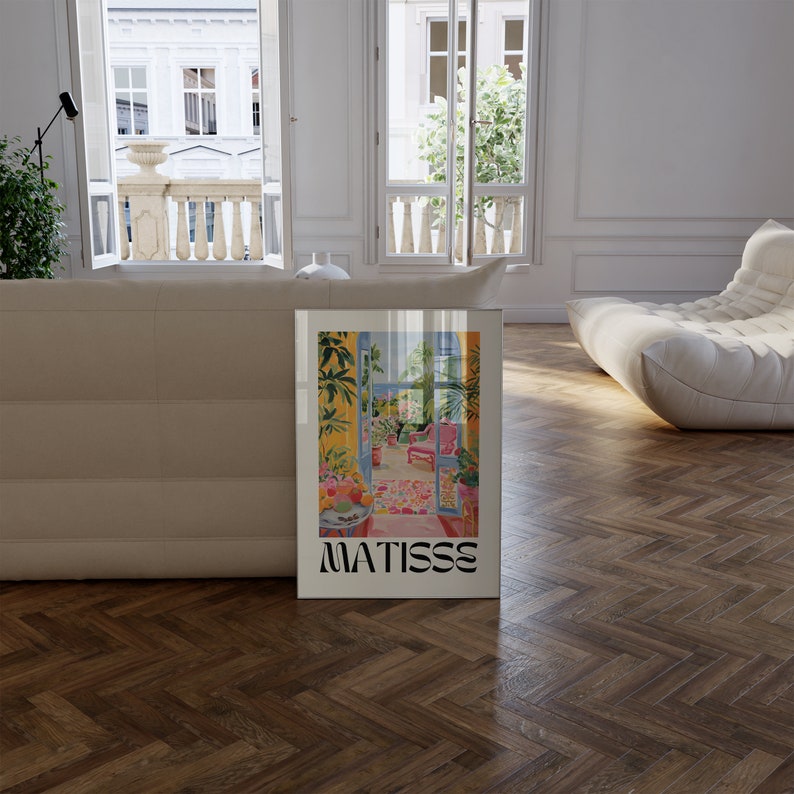 Framed Matisse-inspired poster propped against a beige sofa in a bright room with large open windows and a balcony view. The modern interior features herringbone wood flooring and soft, curved furniture, creating an elegant, airy feel.