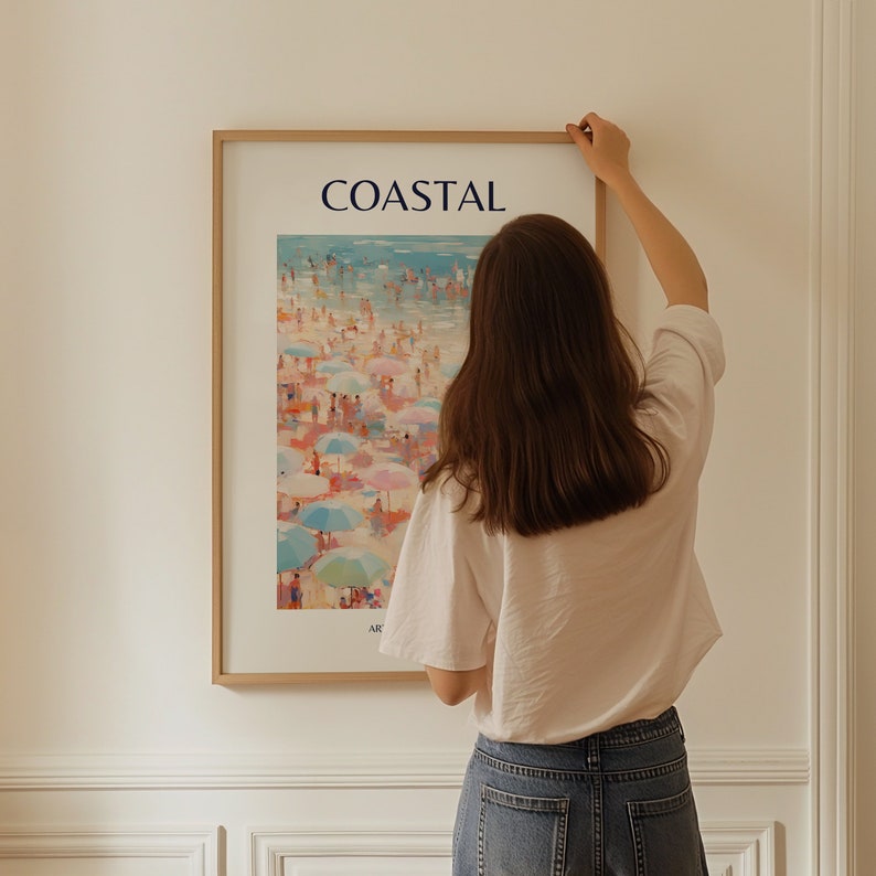 K&ouml;nnte beinhalten: Ein gerahmter Kunstdruck mit dem Titel "COASTAL" zeigt eine Strandszene mit bunten Sonnenschirmen und Figuren. Das Kunstwerk befindet sich in einem hellen Holzrahmen, und eine Person h&auml;ngt den gerahmten Druck an einer wei&szlig;en Wand auf.