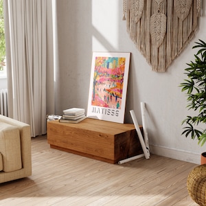 May include: A framed "MATISSE" art print depicting a vibrant street scene, resting on a wooden coffee table. The room features a beige sofa, a macrame wall hanging, and a potted plant. Rolled-up posters are propped against the wall.