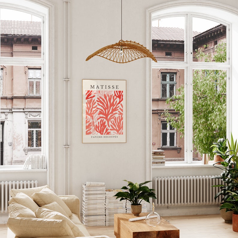 Op de afbeelding: Een ingelijste print van een Matisse-kunstwerk met een rood en roze koraalontwerp met de tekst "MATISSE PAPIERS DECOUPES". De print hangt aan een witte muur in een kamer met grote ramen en een beige bank.