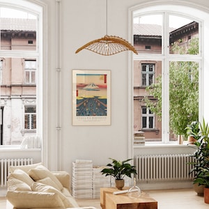 May include: A framed print of a Japanese woodblock print depicting Mount Fuji with a cityscape in the foreground. The print is titled "Utagawa Hiroshige". The print is hanging on a white wall in a living room with a beige couch, a stack of books, and a potted plant.