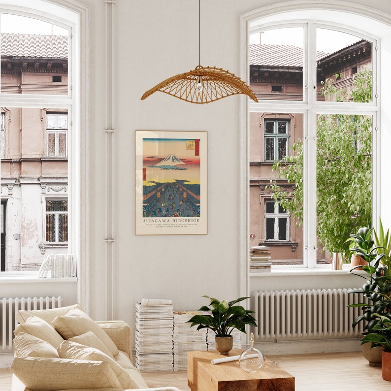 May include: A framed print of a Japanese woodblock print depicting Mount Fuji with a cityscape in the foreground. The print is titled "Utagawa Hiroshige". The print is hanging on a white wall in a living room with a beige couch, a stack of books, and a potted plant.
