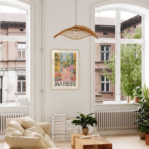 Framed Matisse-inspired poster in a modern living room with large arched windows. The vibrant artwork features a lush garden view with plants and a pink chair. Set against white walls, beige sofa, and greenery, creating a bright, airy feel.