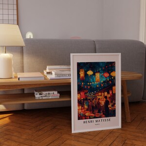 May include: A framed print of a painting by Henri Matisse depicting a street scene in Paris at night. The painting features a vibrant colour palette with red, orange, yellow, and blue hues. The street is lined with buildings and shops, and there are people walking and shopping. The painting is titled "Henri Matisse, Paris".